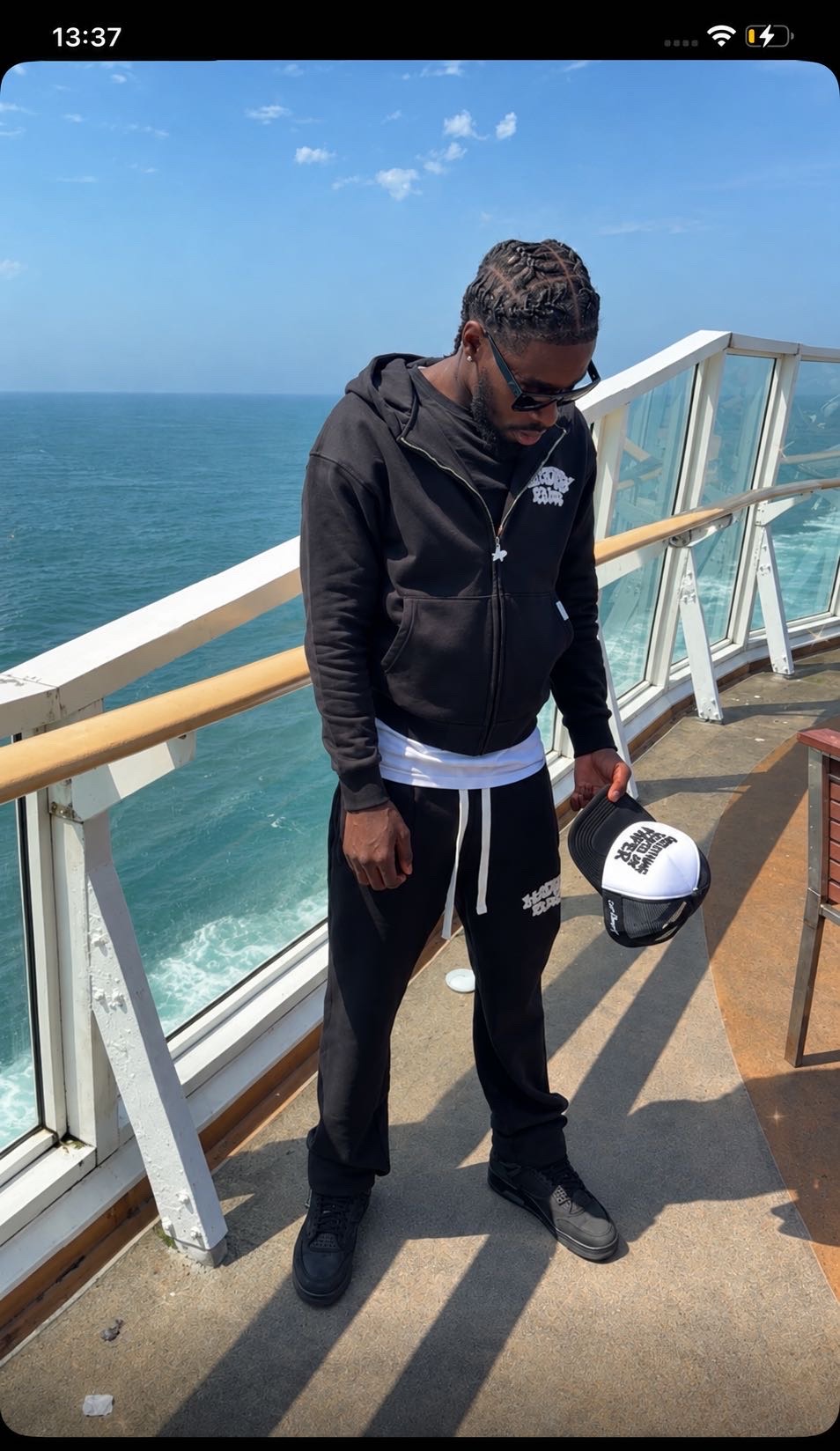 Model on a ferry deck holding a black and white Paper Campaign trucker cap, sea and sky behind him