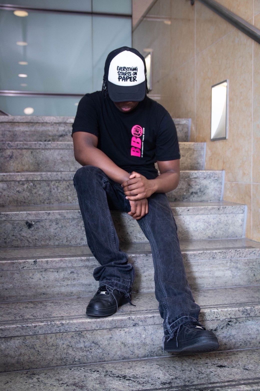 Model sitting on stone steps wearing a black and white 'Everything starts on paper' trucker cap